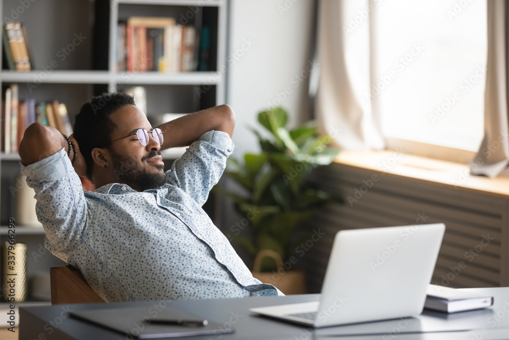 Wall mural Peaceful millennial african american freelance man in glasses chilling on cozy chair, daydreaming alone at home. Sleepy young biracial businessman napping alone at workplace, enjoying break time.