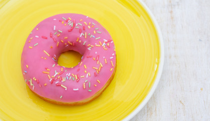 PINK DONUT WITH MULTICOLOR SPRINKLES SERVED IN A YELLOW DISH ON A WHITE WOODEN TABLE