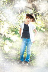 Vertical portrait of 7 years old cute stylish short haired smiling girl in white shirt, blue jeans and brown hat in blooming garden in spring. Short haircut for girl. Allergy season