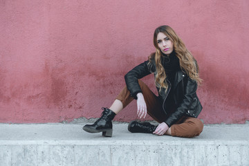 Beautiful millennial female fashion model wearing black leather jacket and brown pants from the fall collection in an urban photoshoot in the street with red wall background