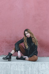 Beautiful millennial female fashion model wearing black leather jacket and brown pants from the fall collection in an urban photoshoot in the street with red wall background