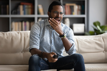 Handsome young african american man in eyeglasses sitting on cozy couch, holding remote controller,...