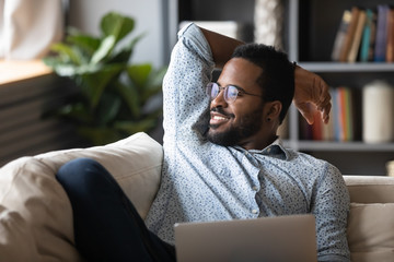 Smiling peaceful handsome african american guy relaxing on comfy sofa with laptop, looking away at...