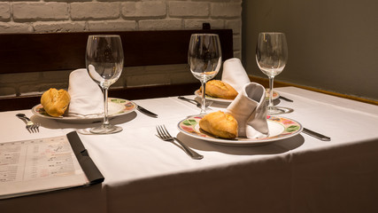 Table laid for three in Spanish restaurant with wine glasses, plates, forks, knives, white table cloths, bread rolls, a menu card.