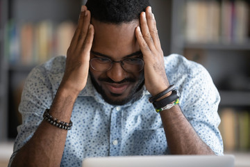 Stressed young biracial businessman holding head in hands, thinking of problem solving, looking at computer monitor. Close up head shot depressed worried african ethnicity guy feeling anxious.