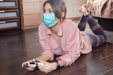 Young attractive woman in hygienic mask plays computer game, laying on the floor of her room....