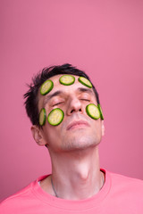 Vertical picture of young man's face full of cucumber slices for face care and beauty. Procedures and fasial treatment. Isolated over pink background.