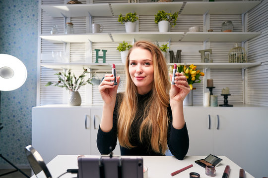 Beauty blogger woman filming daily make-up routine tutorial near camera on tripod. Influencer blonde girl live streaming cosmetics product review in home studio. Vlogger female comparing lipsticks.