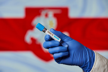 A hand of a doctor in a blue disposable medical glove holding a test tube with a vaccine against coronavirus (COVID-19) against the White-Red-White flag with the emblem Pahonia of Belarus.