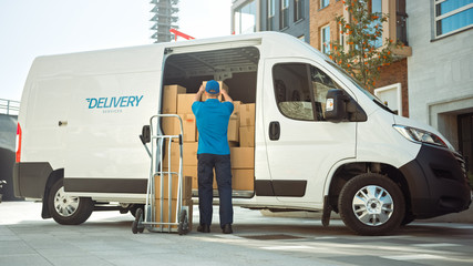 Delivery Man Uses Hand Truck Trolley Full of Cardboard Boxes and Packages, Loads Parcels into Truck / Van. Professional Courier / Loader helping you Move, Delivering Your Purchased Items Efficiently - obrazy, fototapety, plakaty