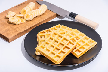 Belgian homemade waffles on a black plate and sliced banana slices on a cutting board with a japanese santoku knife