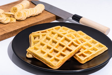 Belgian homemade waffles on a black plate and sliced banana slices on a cutting board with a japanese santoku knife