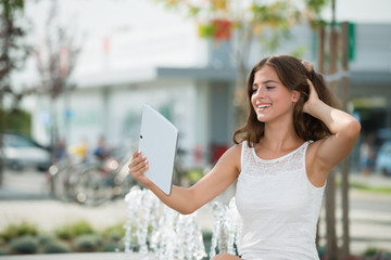 Young teenage girl sitting outdoors, looking at PC tablet, sharing a selfie .Urban scene in the background
