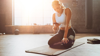 Beautiful and Young Girl Uses Smartphone App During Her Exercise on Fitness Mat. Gorgeous and Athletic Woman Does Mountain Climber Workout in Stylish Hardcore Gym. Shot with Warm Sun Flare