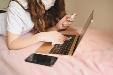 Sick woman in face mask in bed during coronavirus isolation home quarantine cleaning laptop by hand sanitizer, using cotton wool with alcohol. Coronavirus COVID-19 pandemic. Stay home safe concept