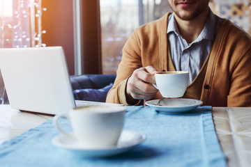 A man sits at a table near a laptop, holds a cup of coffee, close-up
