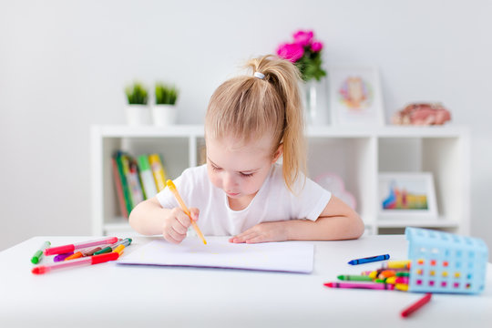 Blonde happy little girl with ponytale drawing and writing lsitting by the white table in light roon. Preschool education, early learning