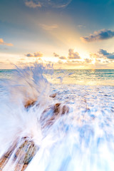 Detail view of rock on the beach, placed in the sand, with big water wave from the sea. Beautiful sunset above ocean with soft colors and clouds on sky. Concept of tropical vacation, relaxation.