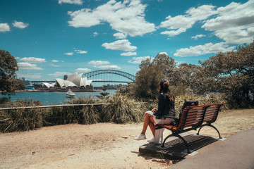 Naklejka premium Woman with Sydney Opera House & Harbour Bridge. Tourist looking at attraction, with river water. Blue sky tourism shot. Boats on river. Famous landmark.