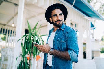 Pleased ethnic hipster using smartphone outside cafe