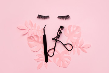 Black false lashes strips and tweezers on pink background with paper palm leaves