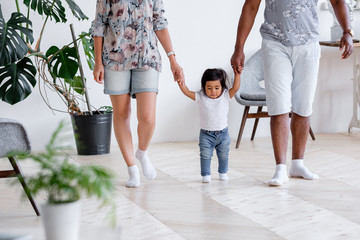 Positive little one and half year mixed-time girl walks around house and takes her first steps holding hands of father and mother in cozy home. Concept of care of love and support for next generation