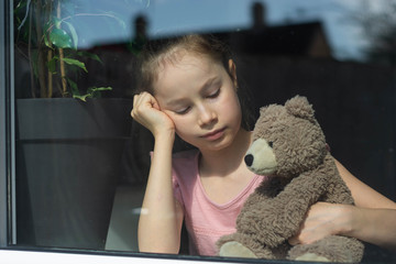 Sad little girl with her teddy bear at the window. homeschooling education.