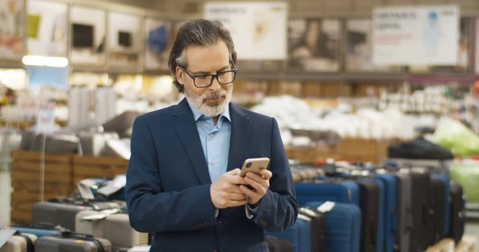 Handsome Caucasian man texting message on mobile phone in supermarket. Stylish businessman scrolling and tapping on cellphone at suitcases in shop. Male shopper with smartphone in hypermarket.