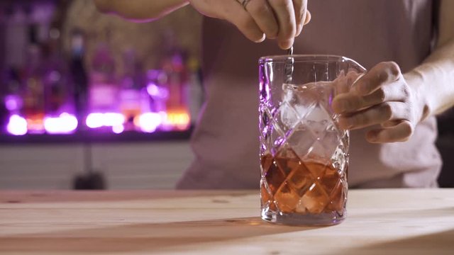 Male Bartender's hand Mixologist Combining Ingredients and Making Refreshing Negroni Classic Cocktail in Trendy Bar. Close-up of Barman Stirs Ice Cubes with Small Spoon. Purple light