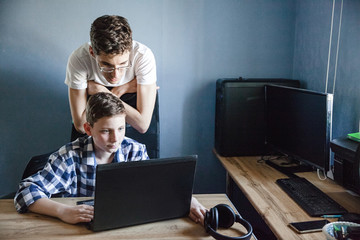 Brothers in the process of distance learning. The younger brother sits at the table and looks at his laptop. The older brother stands behind him and controls the process