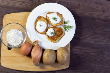flat lay on Potato fritters dranik with sour cream on white plate and ingredients for cooking (potato, onion, egg, powder) on brown wooden background with copy space