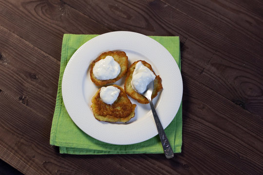 Potato fritters dranik with sour cream and spoon on white plate on brown wooden background
