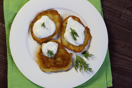 Potato fritters dranik with sour cream and dill on white plate on table cloth on brown wooden background