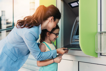 Fototapeta na wymiar Young mother elegantly dressed with her daughter using ATM machine on city street.