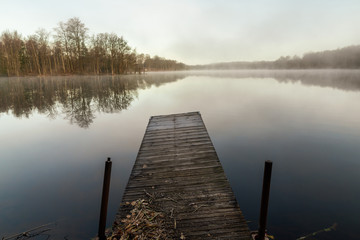 A jetty by a mirrored lake on an early morning with fog easing