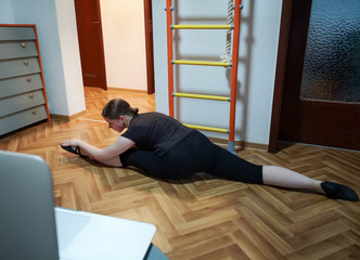 Blurred shot of teenage girl doing gymnastics and sitting in the splits on floor at home remotely with coach via online streaming platform on laptop.Home space with sports ladder,rope is in background