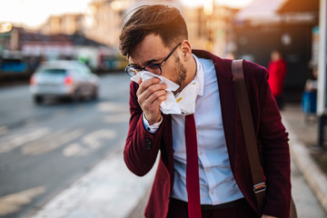 Young businessman sneezing while standing alone on empty street and waiting for bus or taxi. Virus pandemic or pollution concept. © hedgehog94