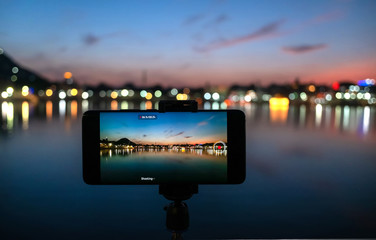 A phone on tripod recording the sunset over a lake (focus on phone device and tripod, city  blurred)