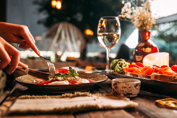 Side view appetizing italian cuisine dishes caprese salad with tomatoes with mozzarella cheese and prosciutto bruschettas, hands with holding cutlery, horizontal format