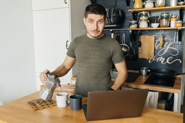 positive young man remotely pours himself coffee in the interior of the home kitchen and works with a laptop on the table. Morning, quarantine, self-isolation.