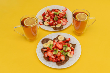 Serving for two. Fresh pancakes with chopped fruits, strawberries, bananas, kiwi and honey on light plates. Two cups of black tea with lemon. Healthy food, gluten free. Copy space. Yellow background