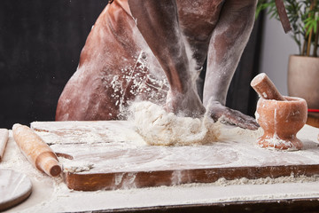 Dark skin baker man kneads Dough in the kitchen. White flour flies in air on black background, pastry chef claps hands and prepares yeast dough for pizza pasta.