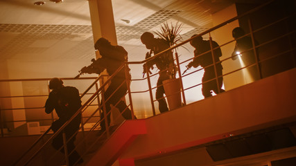 Masked Squad of Armed SWAT Police Officers Run Down the Stairs from a Second Floor in a Dark Office Building. Soldiers with Rifles and Flashlights Move Forwards, Cover Surroundings. Warm Color Grading