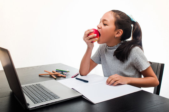 Little girl eating an apple while studying at home following online classes on the laptop. Concept of studying at home on white background.