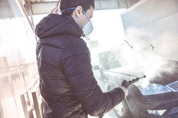 washing car by water pressure at self-service station. A man with a respirator on his face protects...