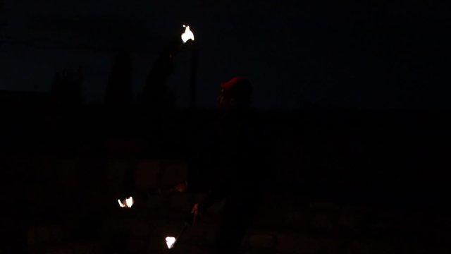 Professional juggler performing juggling tricks with 3 fire clubs, lighting the outdoor dark night scene. Slow motion medium shot.