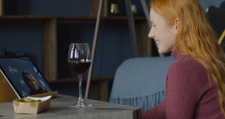 Attractive mid-30s Caucasian female clinking glasses while chatting with a friend or partner online in video chat, using a tablet