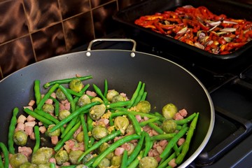 Green beans and brussel sprouts with bacon bits in a deep pan on an oven top together with a roasting dish with red peppers and onions in the background against a copper tiled backsplash.