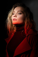 Beautiful girl wearing a casual trendy trousers, turtleneck glasses and raincoat posing on a dark background in the shadows and red light. Fashionable, advertising, lifestyle and commercial design.