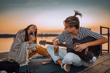 Happy young couple drinking beer, playing a guitar and singing while sitting on a wooden pontoon...